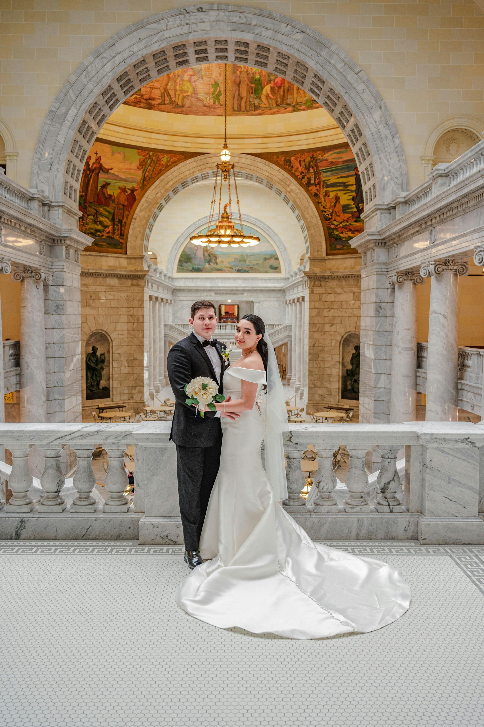 Bride and groom posing in an ornate arched hall with a chandelier and marble balustrade