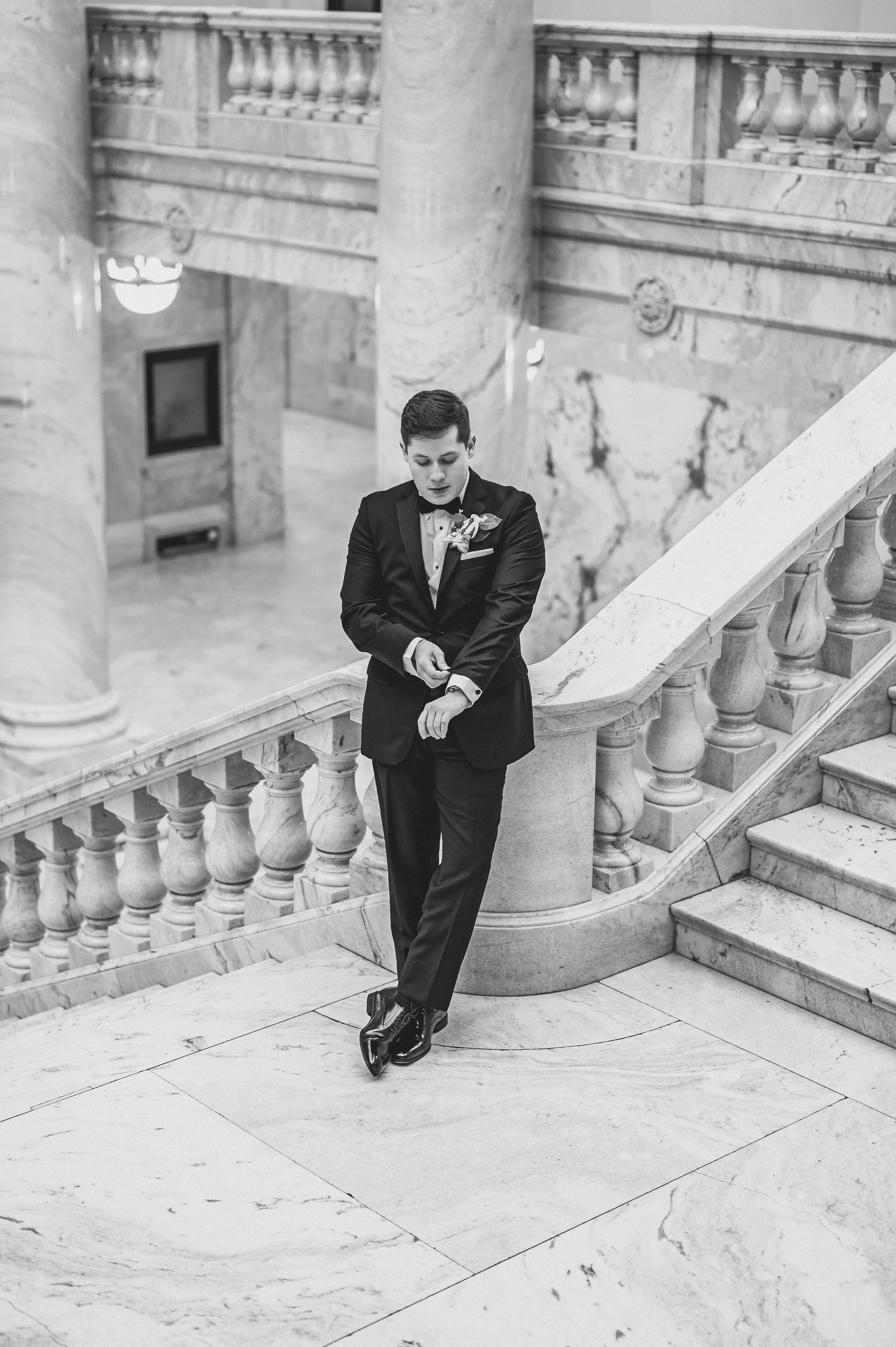 Man in a tuxedo leans on a grand staircase in a marble building.
