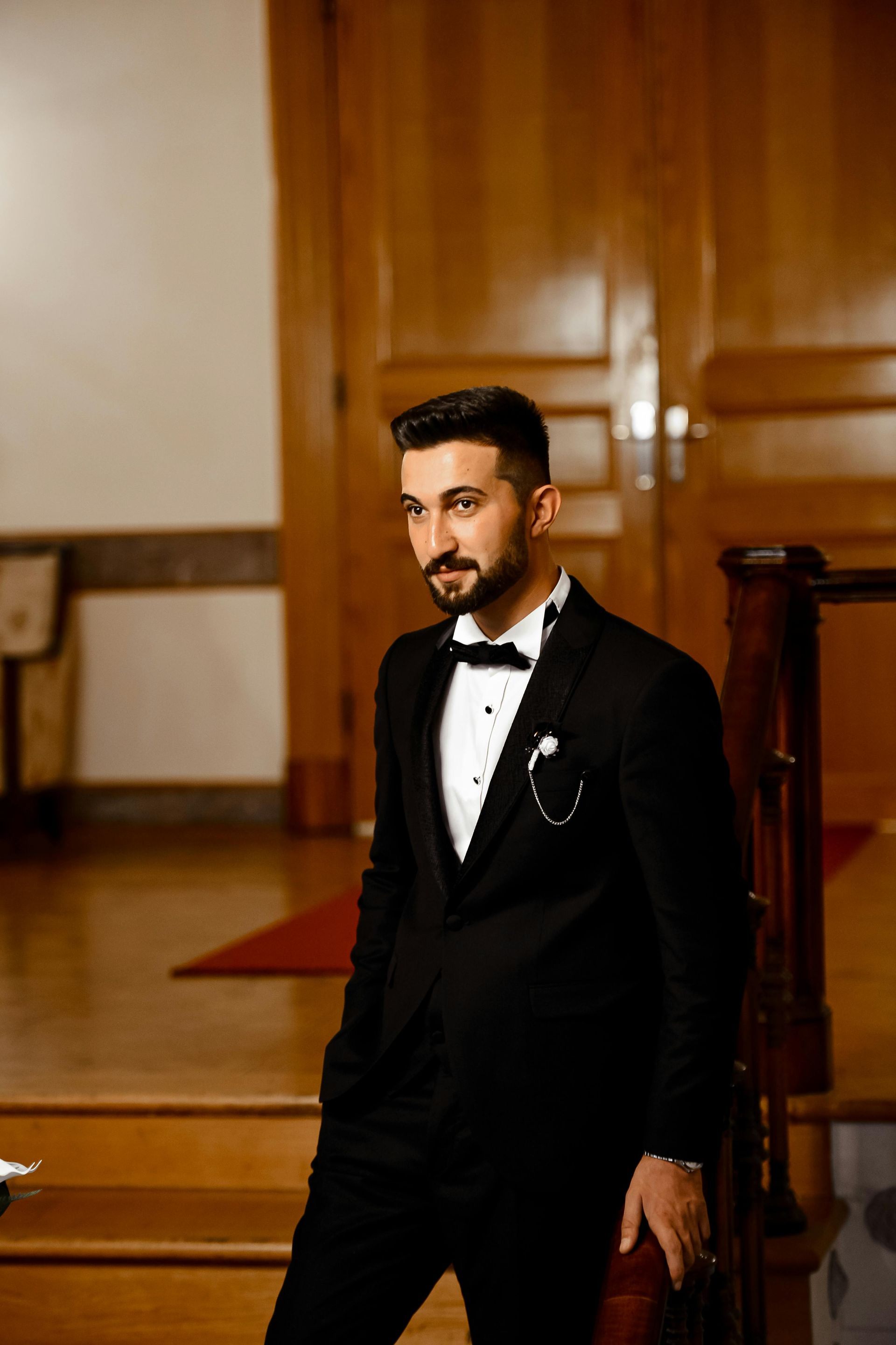 Man in a black tuxedo standing on a staircase in a formal indoor setting