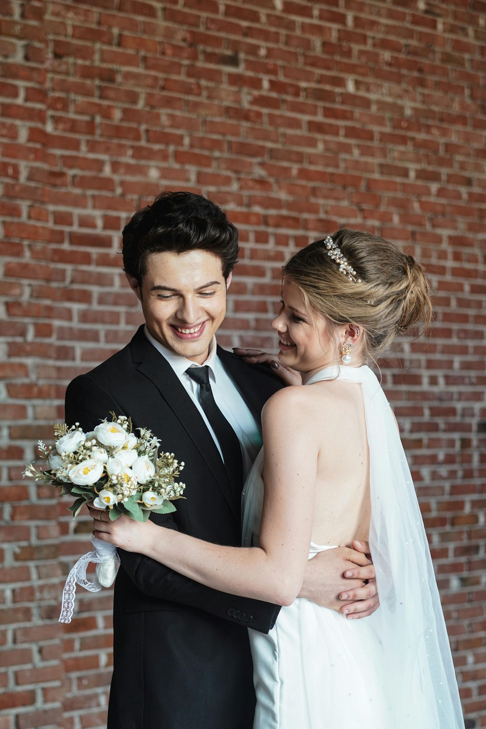 Bride and groom embracing and smiling with bouquet in front of a brick wall