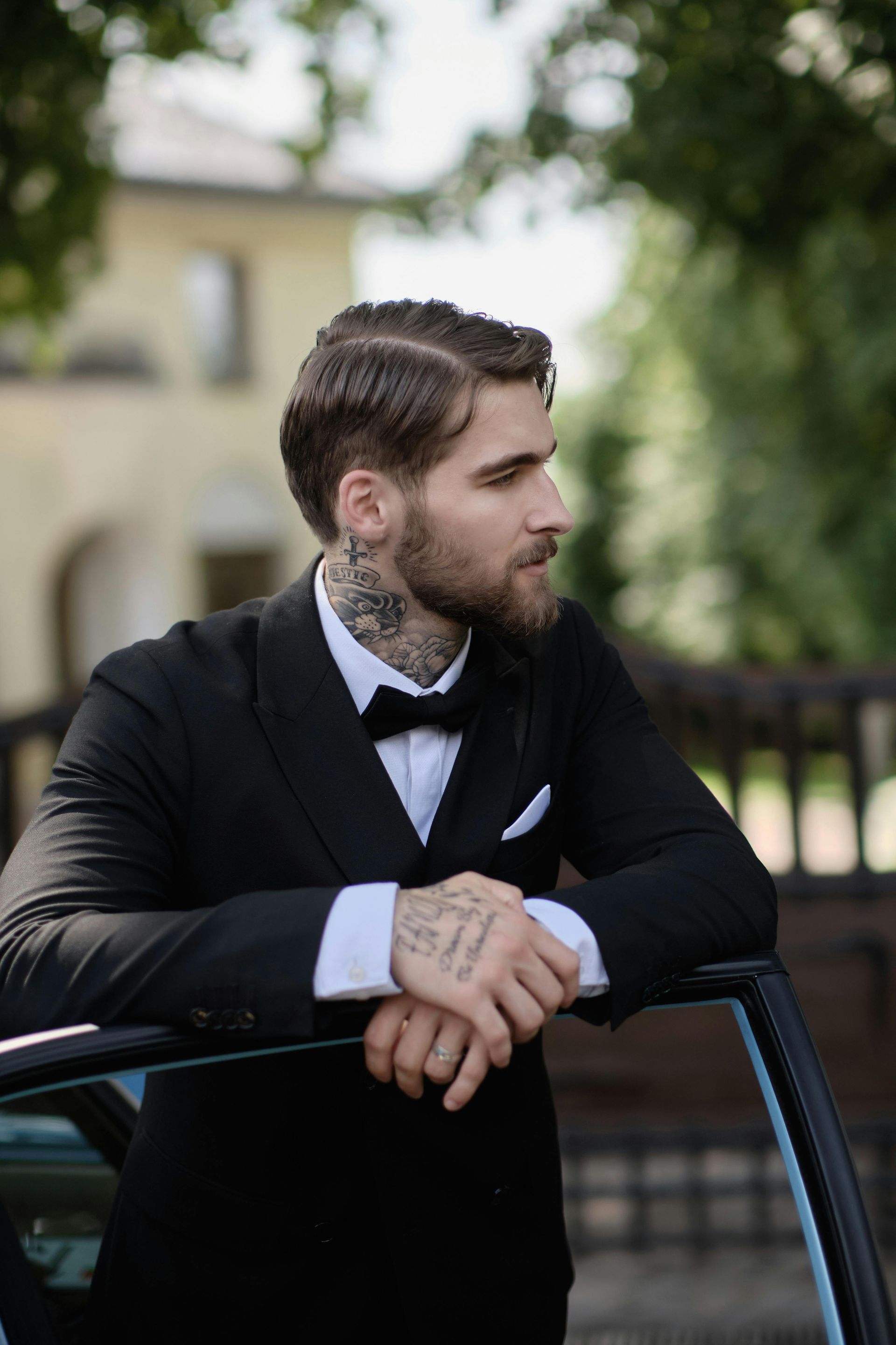 Man in a black tuxedo leaning on a car door, looking away outdoors.