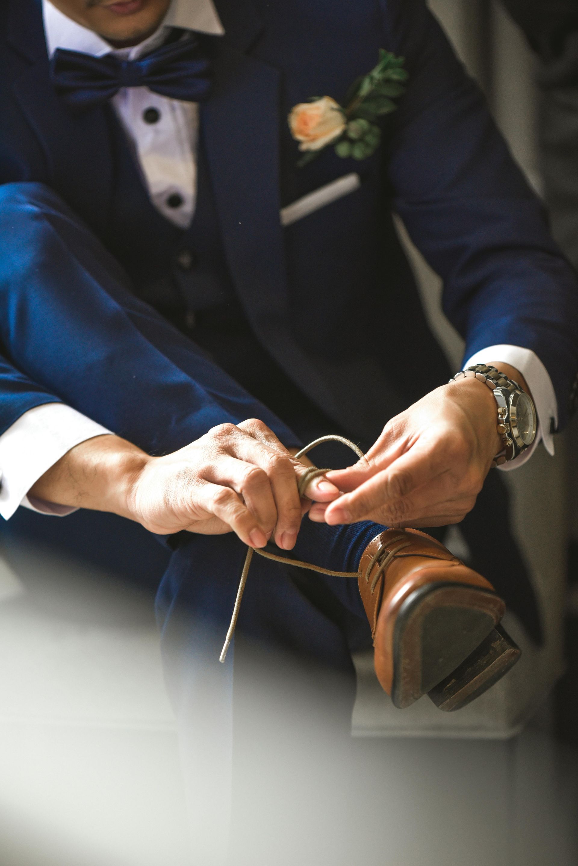 Man in a navy suit tying a brown dress shoe, likely during wedding preparation