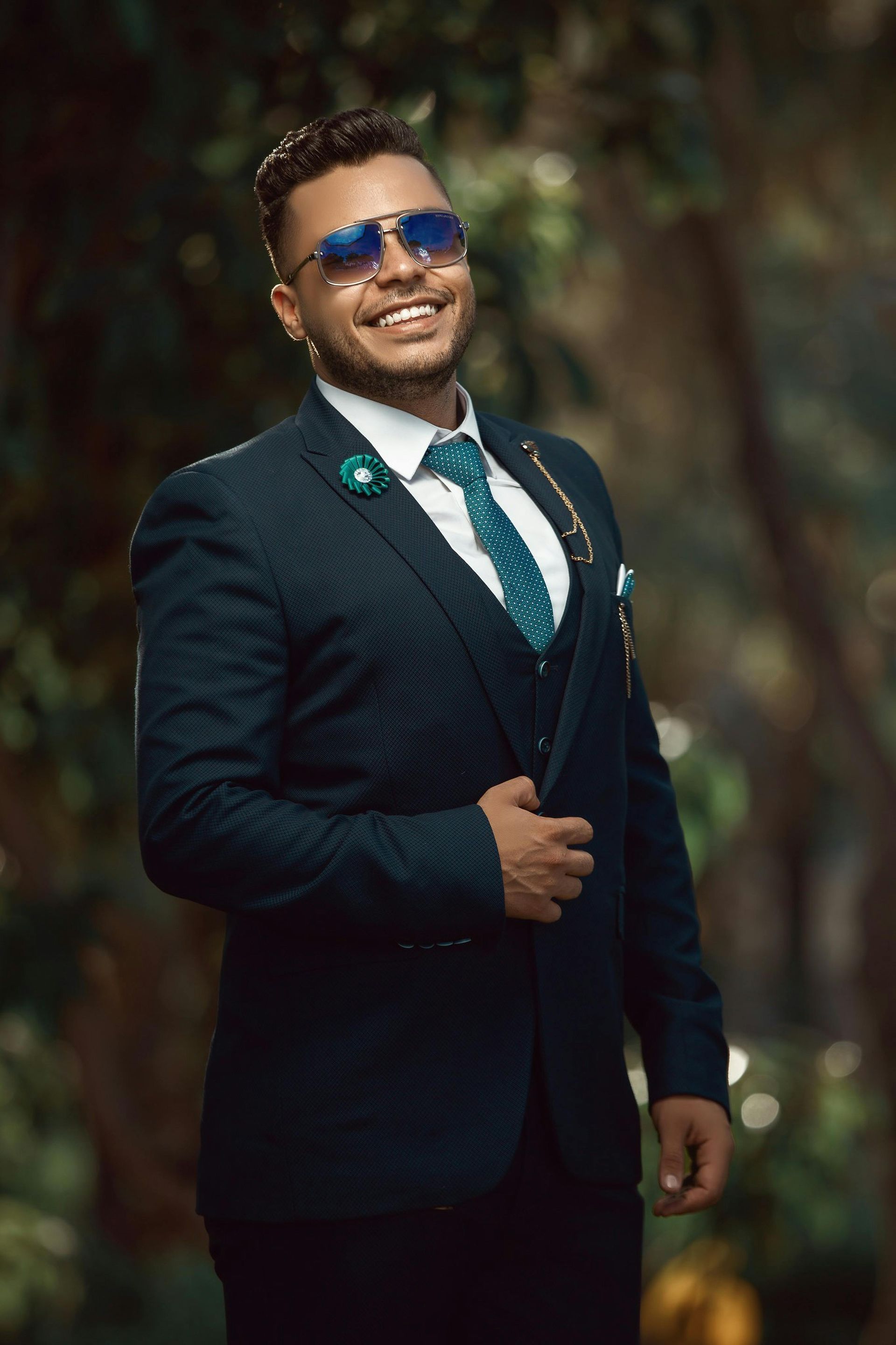 Man in a dark green suit and sunglasses smiling outdoors against a blurred background.