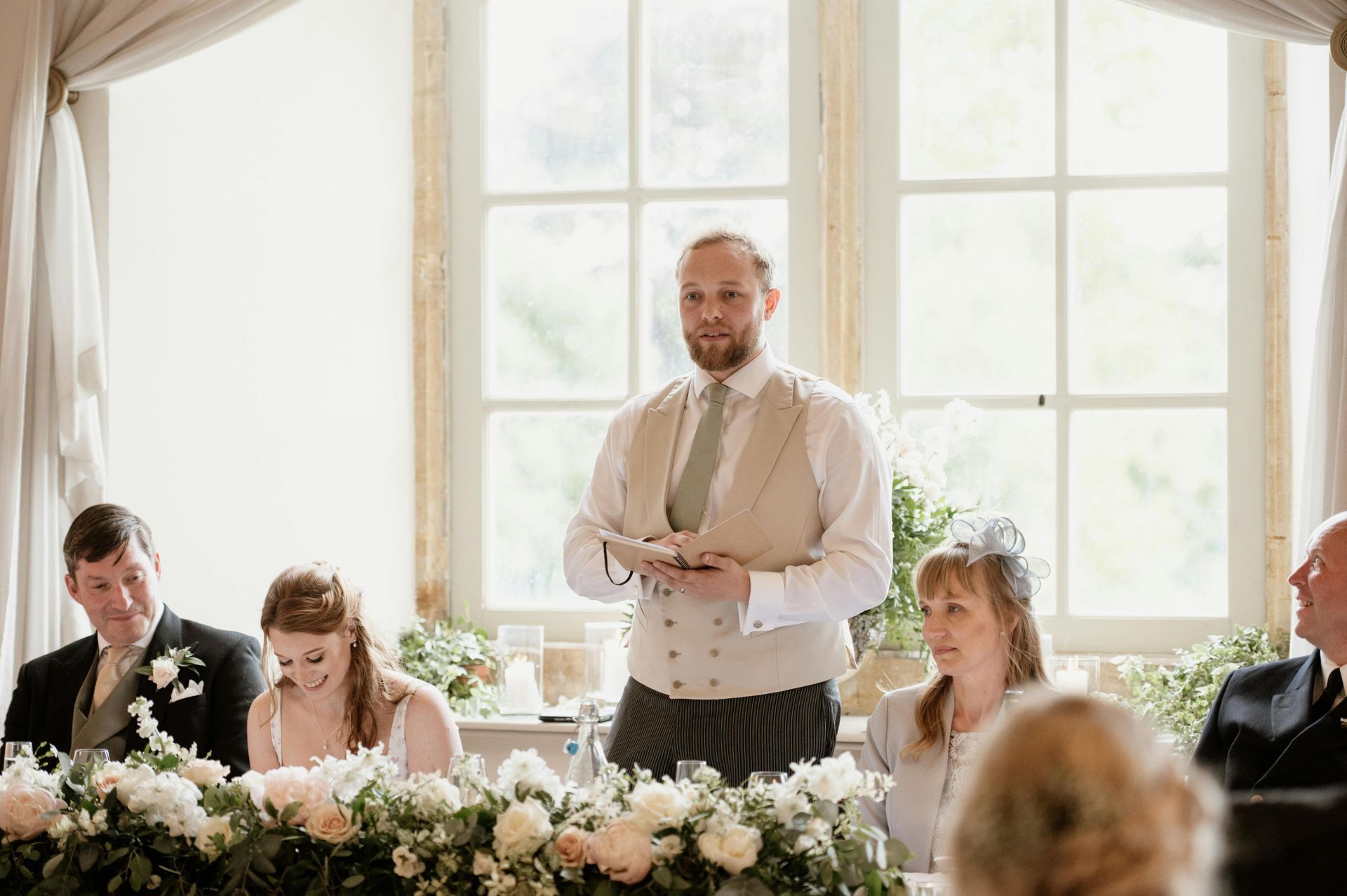 Man giving a speech at a wedding reception table decorated with flowers, guests seated beside him in bright window light