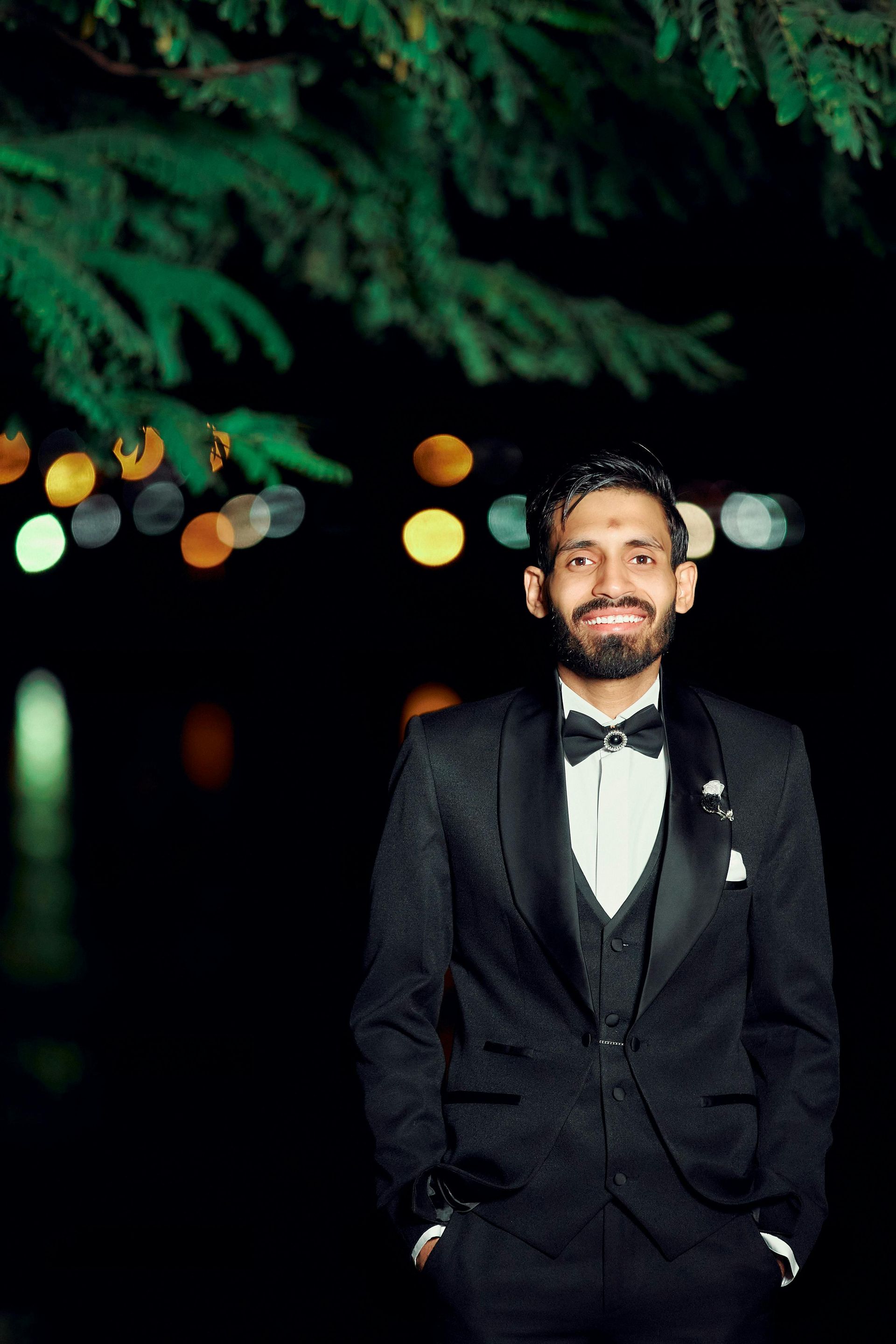 Man in a dark suit and bow tie smiling outdoors at night, with blurred lights and greenery behind him