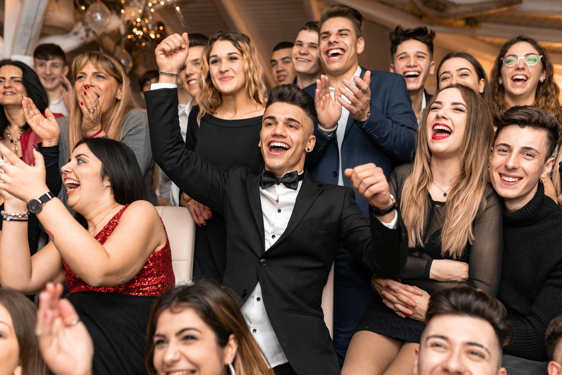 Crowd of people in formal wear cheering and smiling at an event, with one man in a black tuxedo raising his arms.
