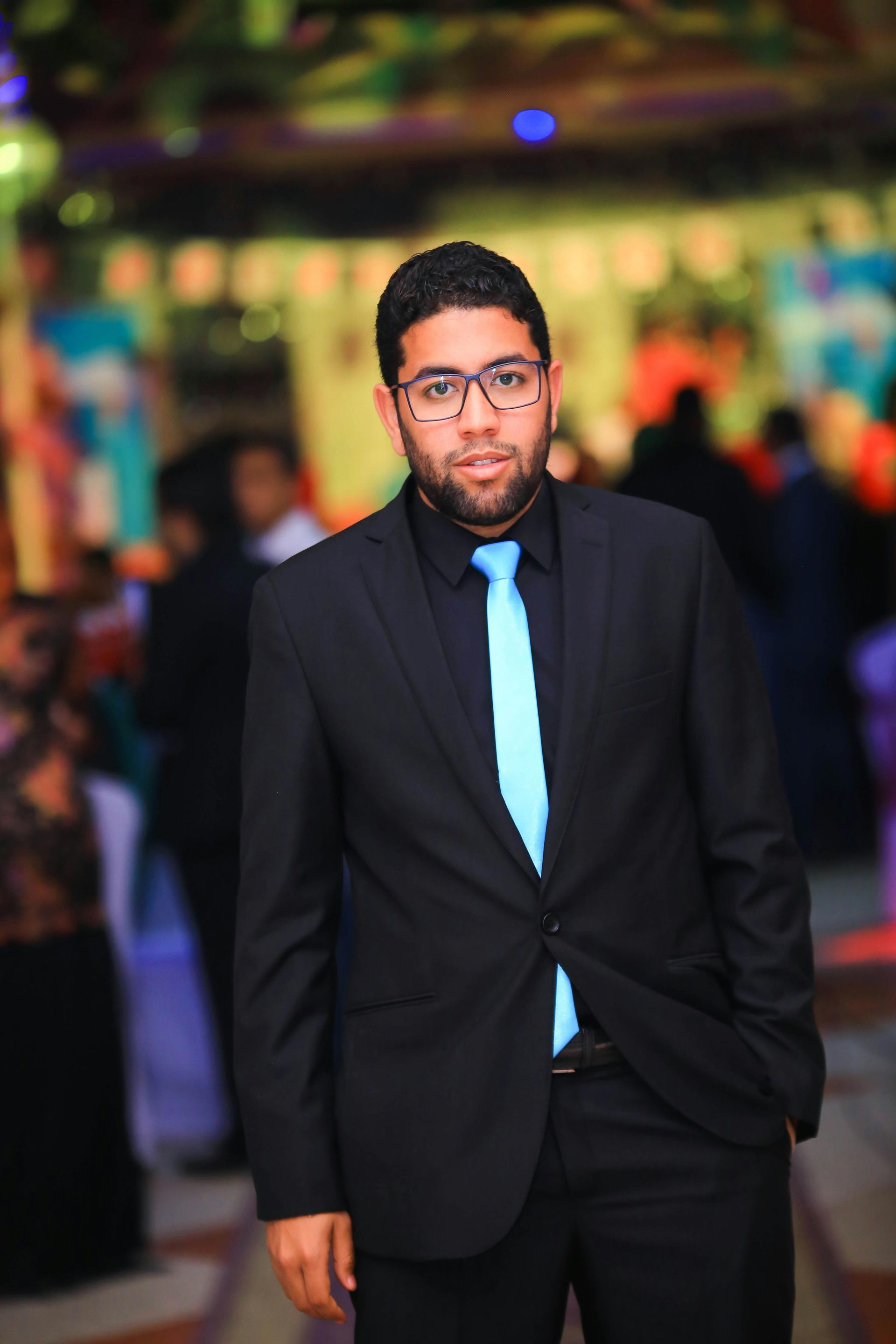 Man in a black suit with a light blue tie standing outdoors against a blurred colorful background