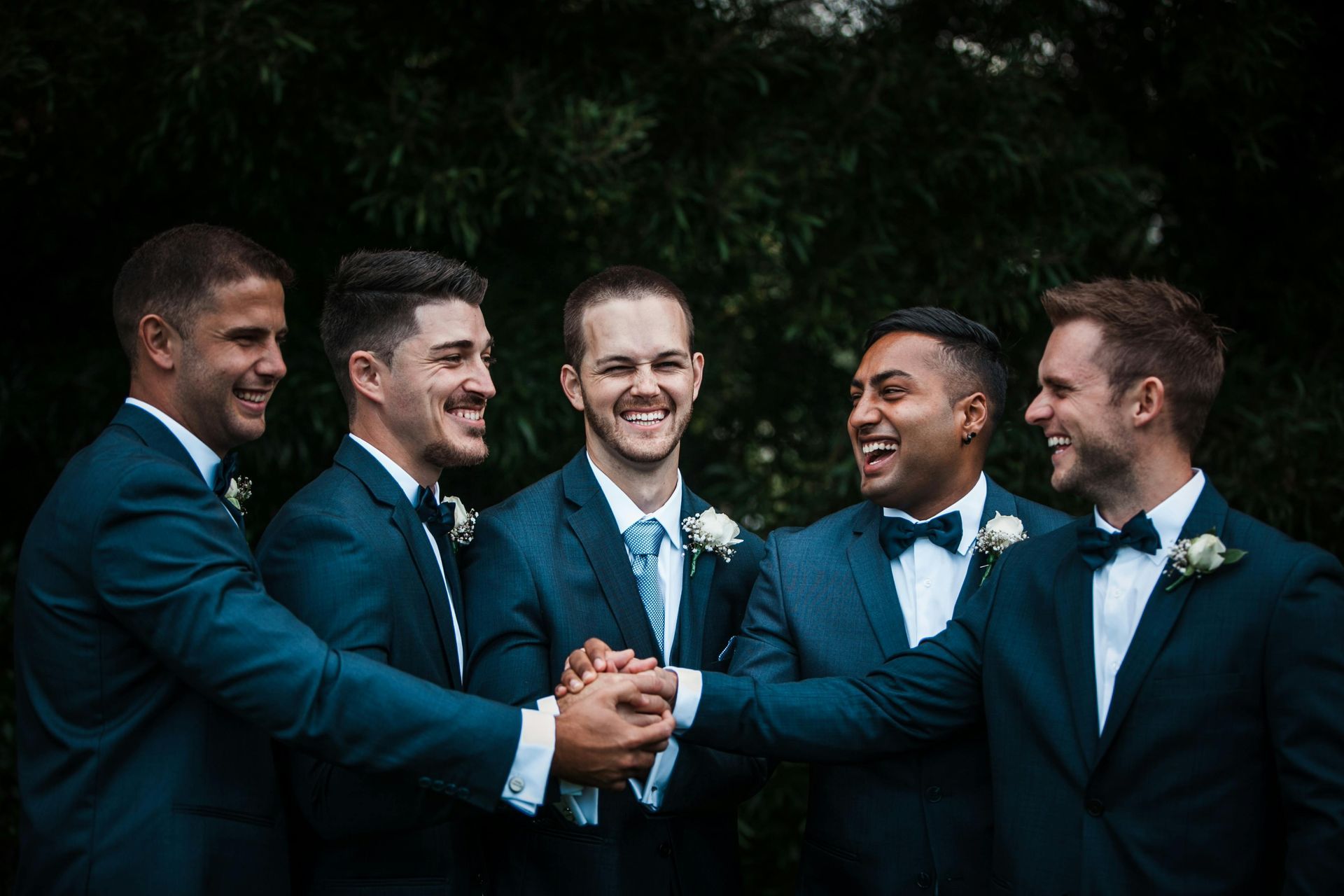 Five men in teal suits smiling and stacking hands outdoors at a wedding