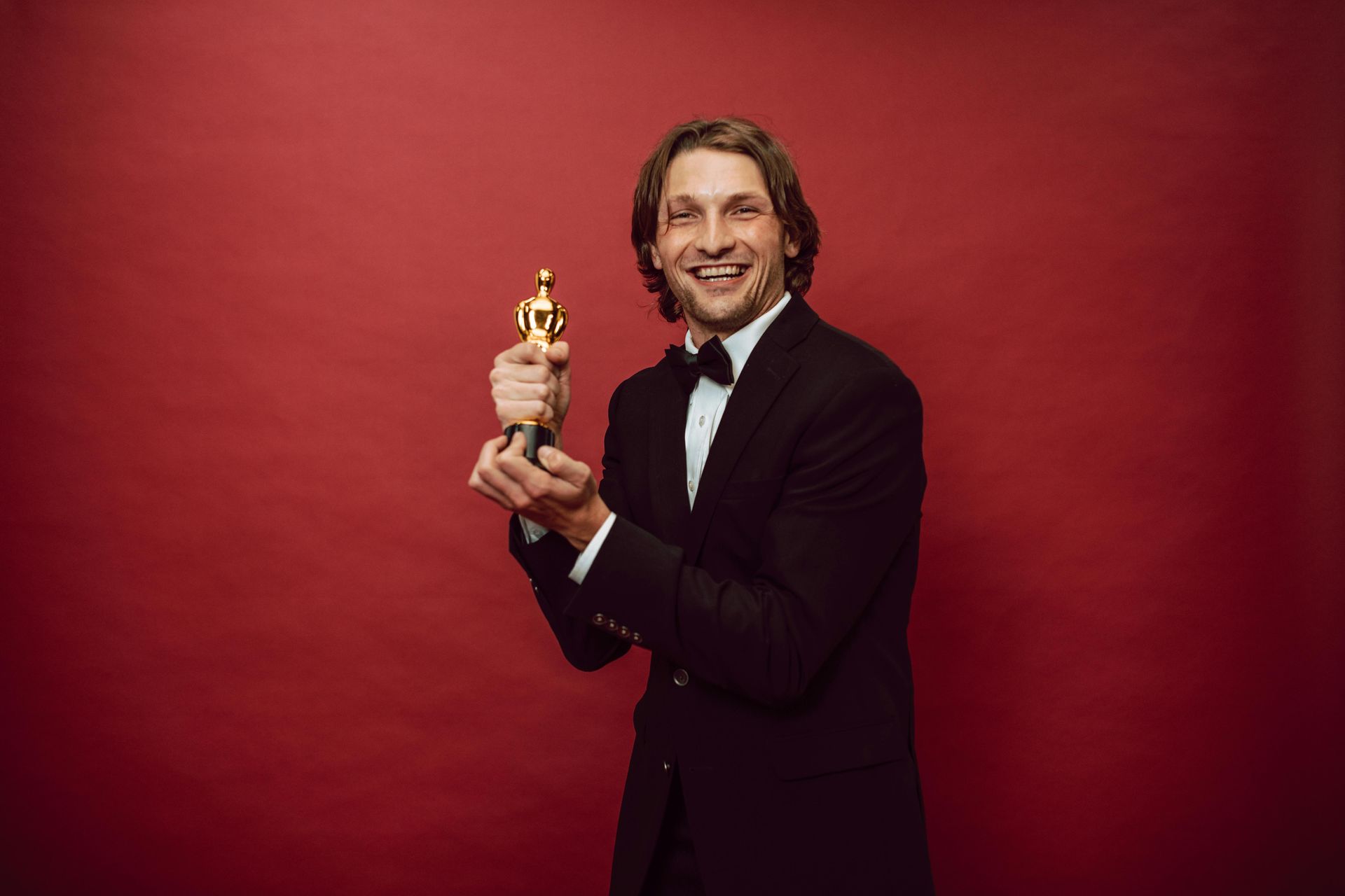 Man in a black tuxedo smiling and holding an Oscar statuette against a red backdrop
