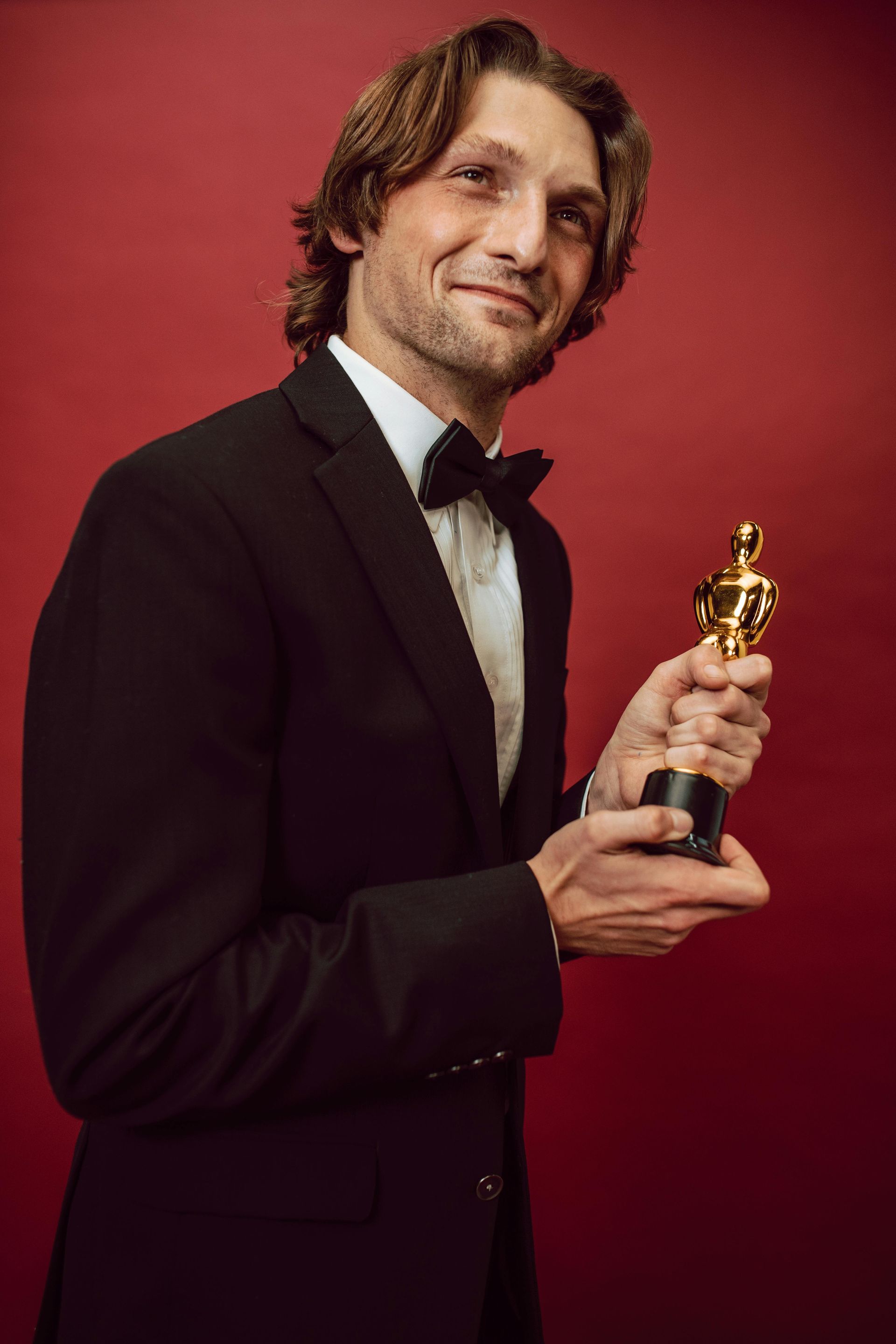 Man in a black tuxedo holding an Oscar statuette against a red backdrop