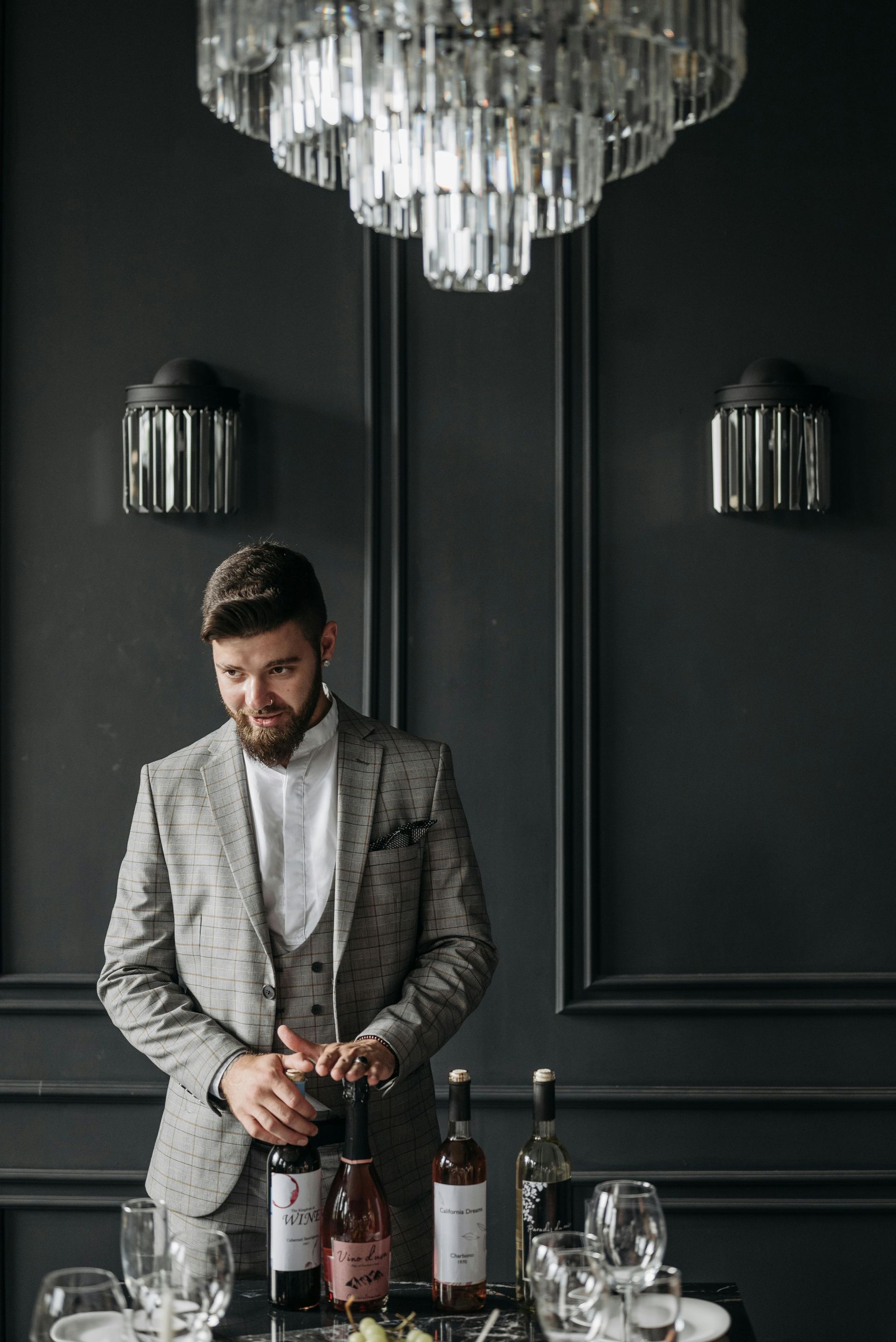 Man in a gray suit preparing drinks behind a bar with crystal chandelier and dark paneled walls