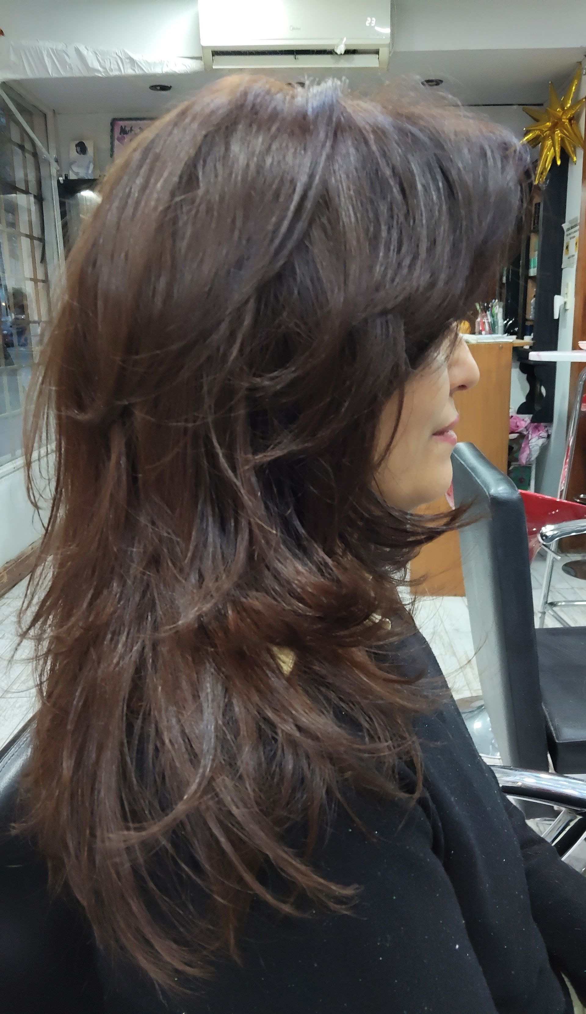 Una peluquera peina el cabello rizado de una clienta en un salón luminoso con paredes y espejos rosas.