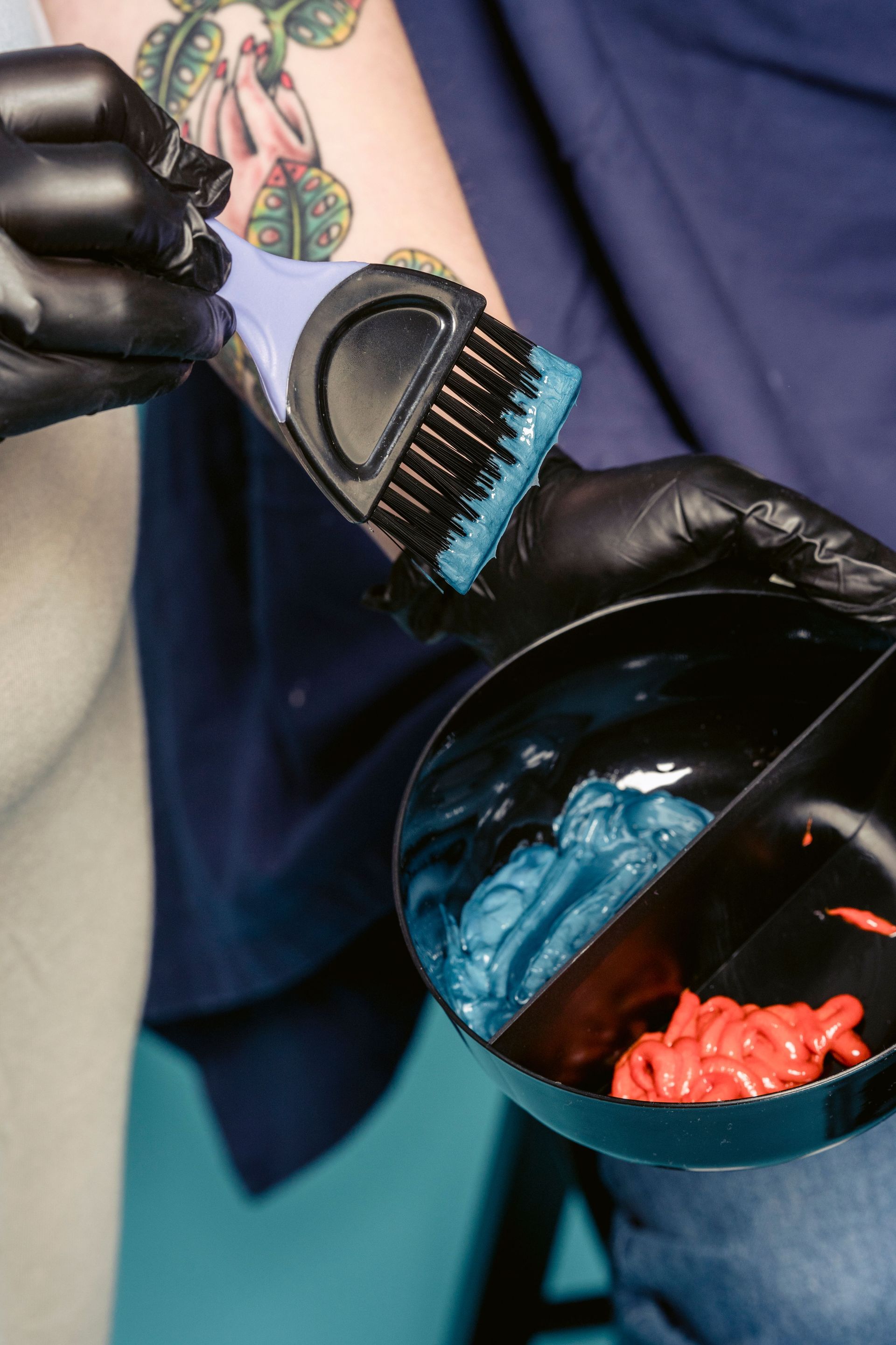 Una mano enguantada tiñe el cabello con un pincel junto a un recipiente con tinte azul y rojo.