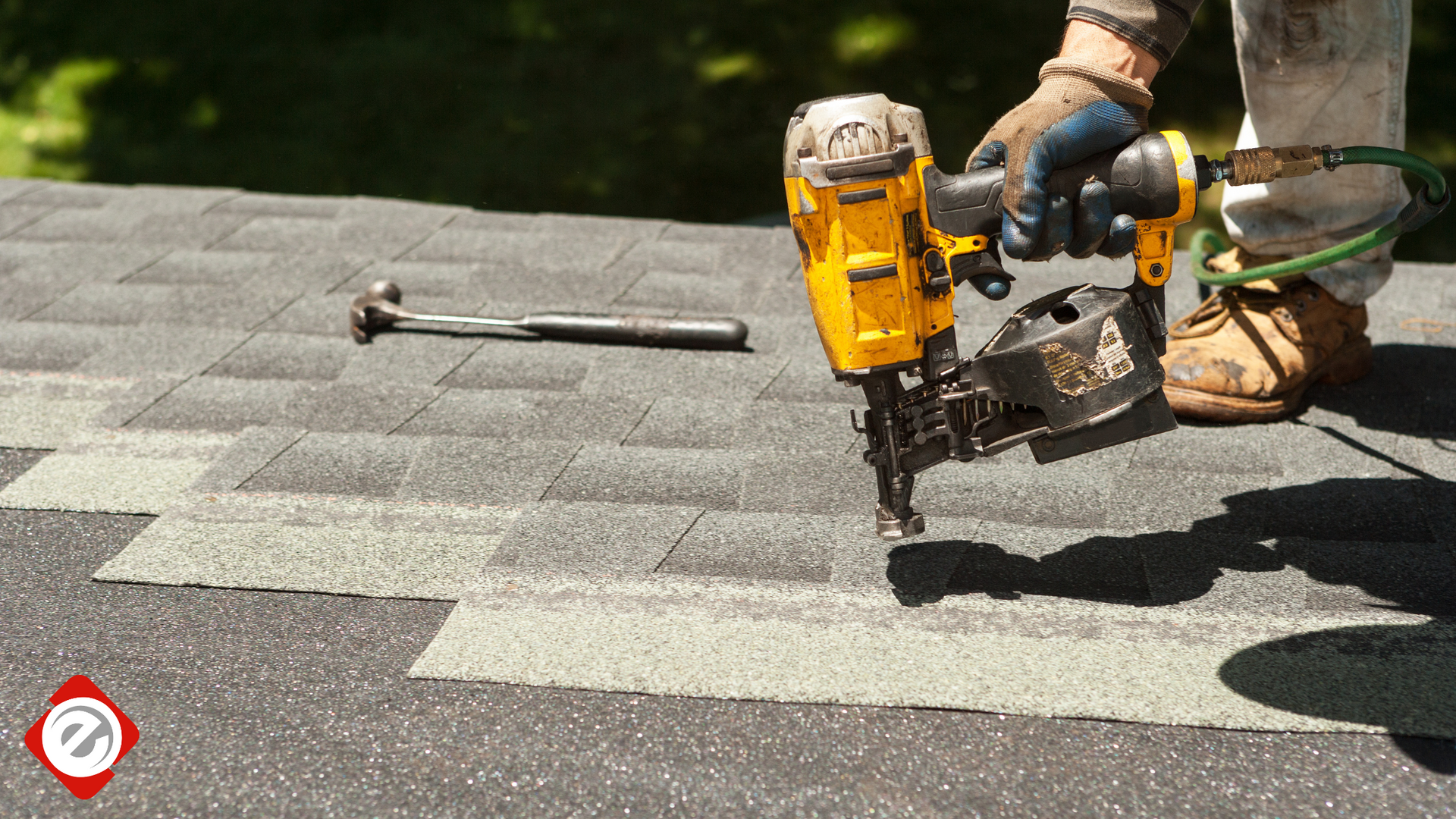 Roofer using a nail gun on a dark shingle roof. A hammer lies beside the gun.