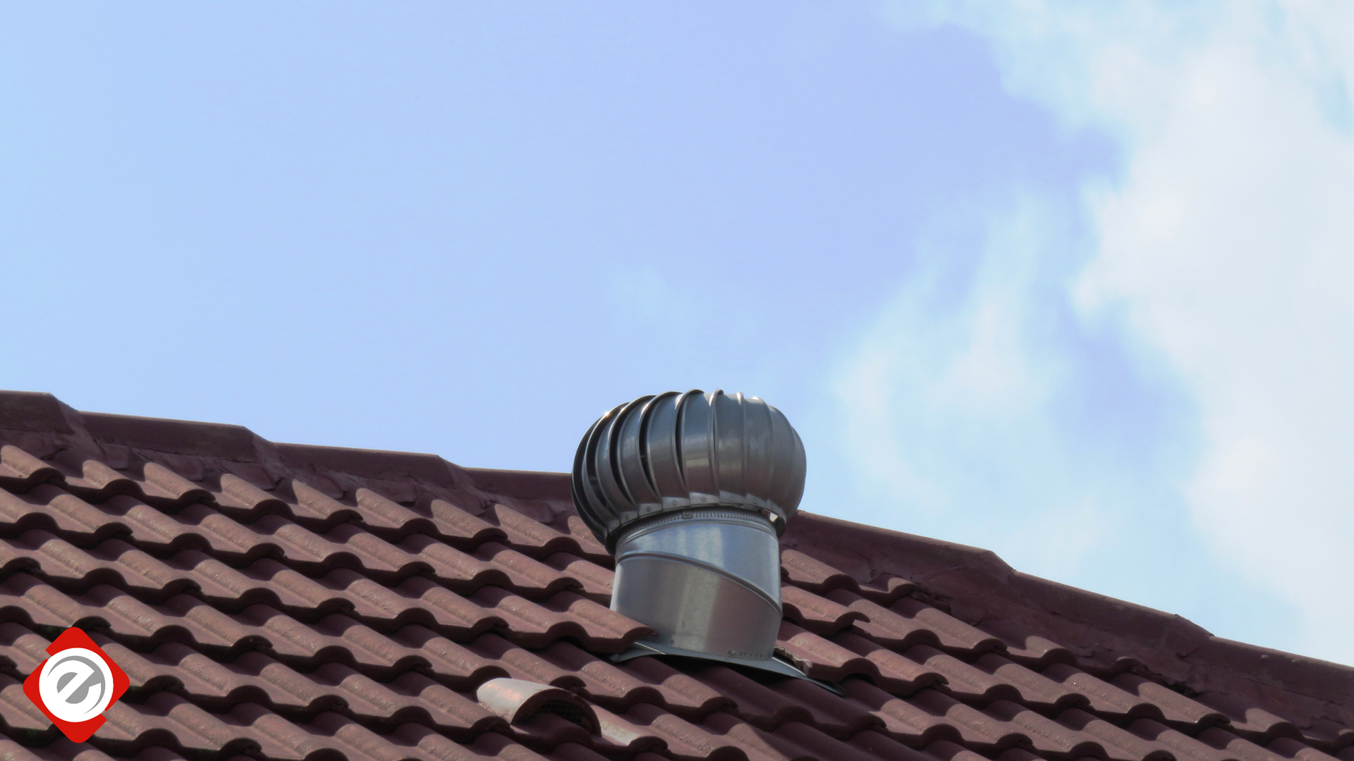 Metal roof turbine ventilator, brown roof tiles, against blue sky.