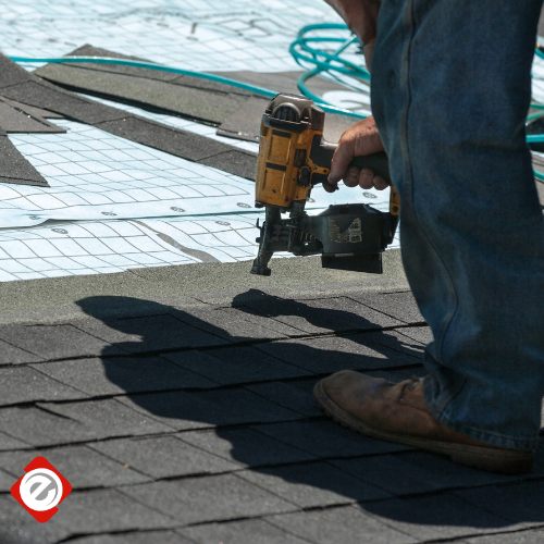 Roofer using a nail gun to install shingles. Wearing jeans and work boots on a sunny day.
