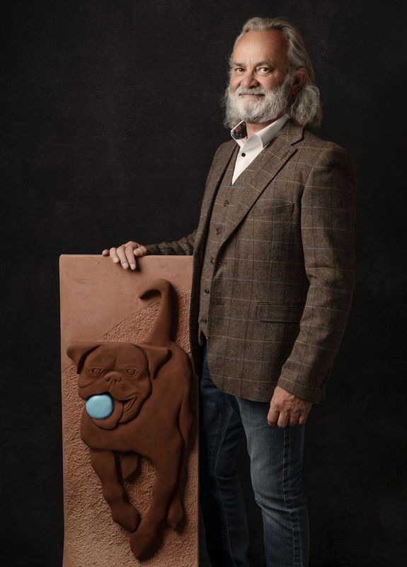 A man with a beard is standing next to a sculpture of a dog with a ball in its mouth.
