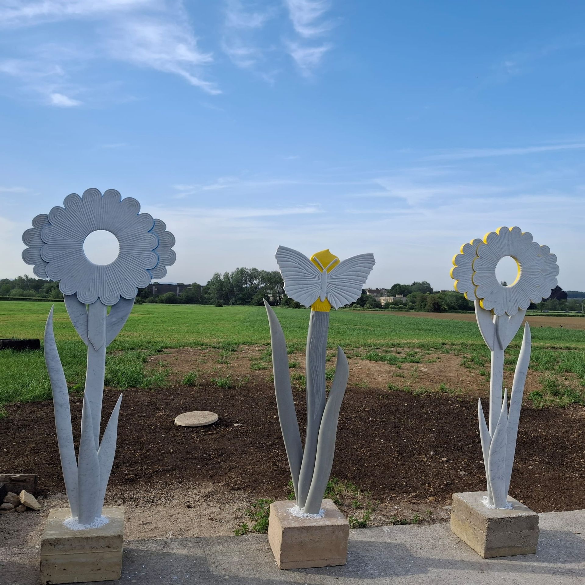 Three metal flower sculptures on stone bases in a grassy field under a blue sky.