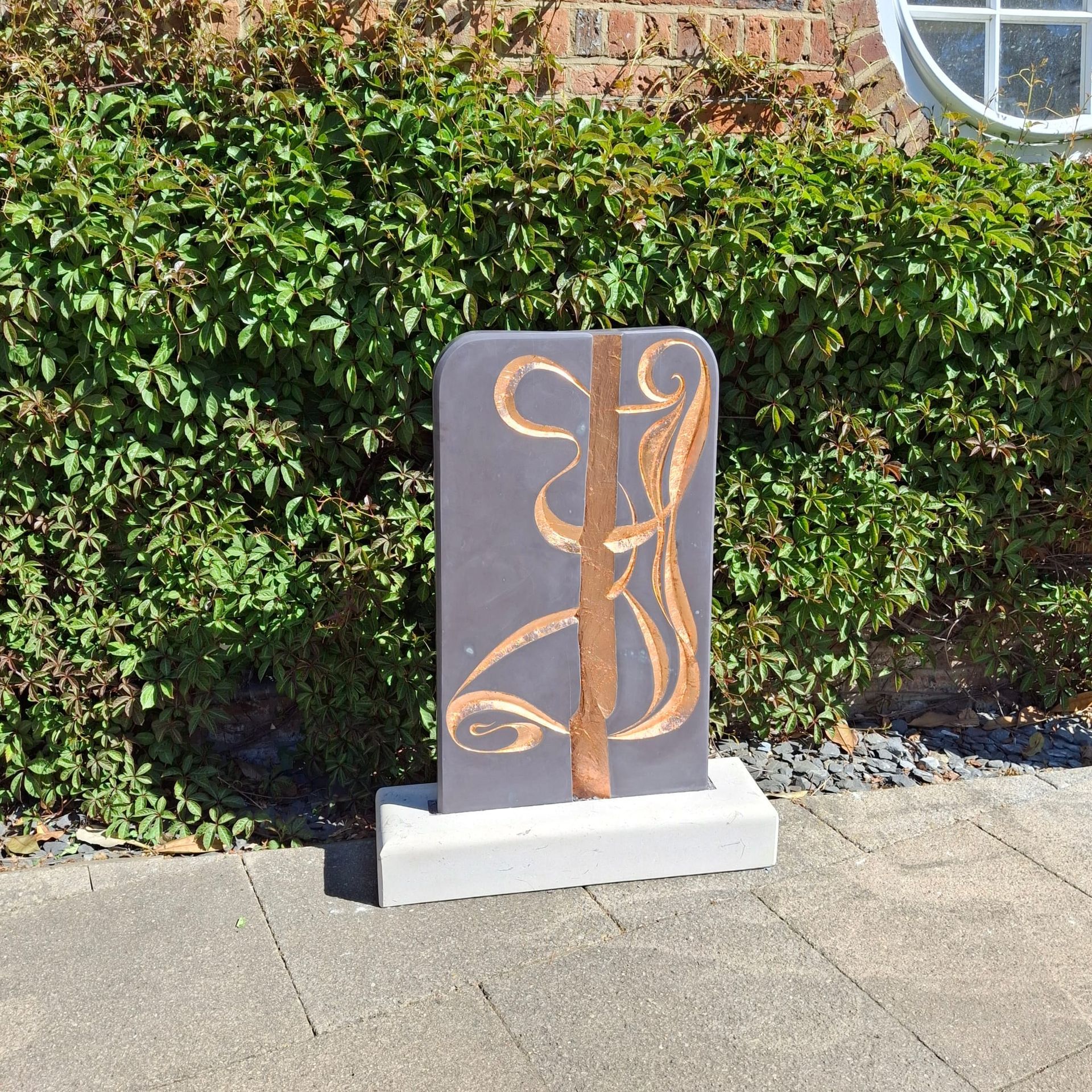 Abstract bronze sculpture on a white base beside a hedge and brick wall outdoors