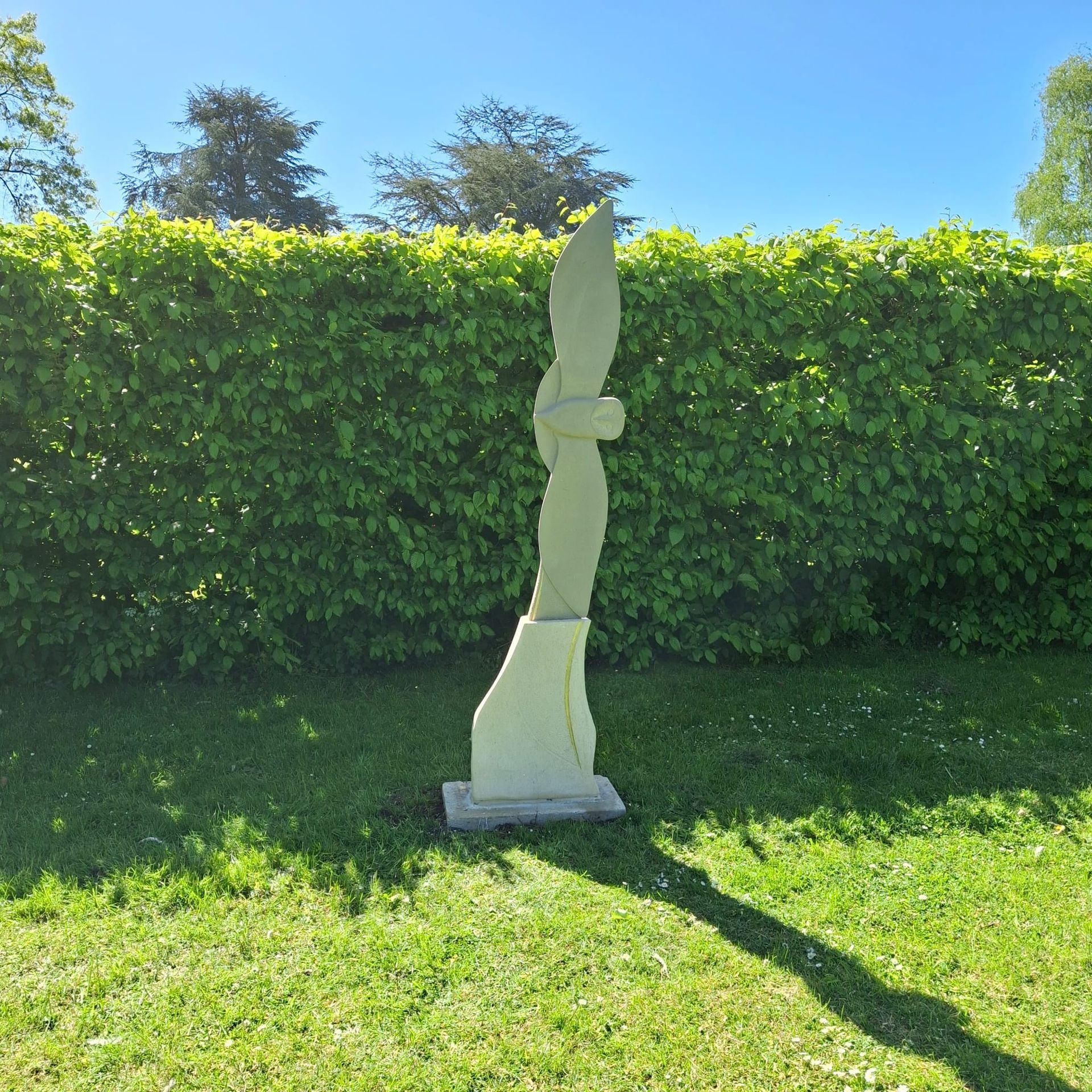 Tall abstract stone sculpture on a grassy lawn in front of a green hedge under a blue sky.