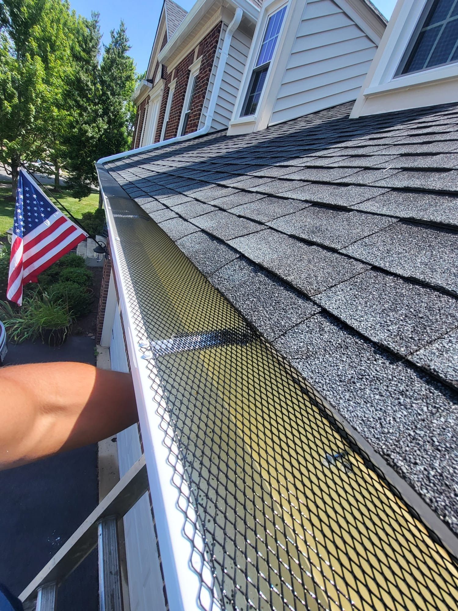 Gutter guard installed along a shingled roof beside a house with an American flag on the lawn.