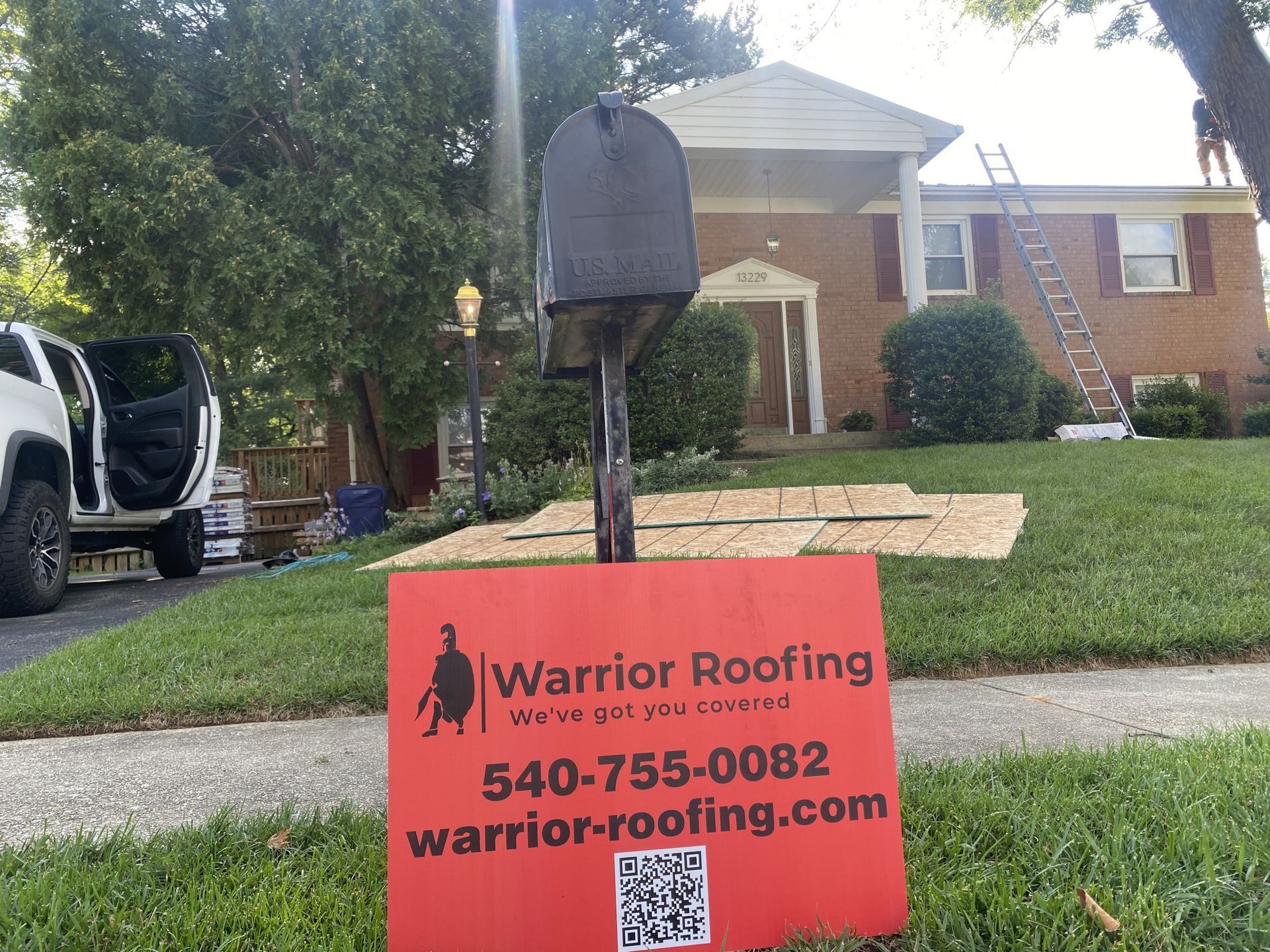 Warrior Roofing yard sign in front of a suburban brick house with a mailbox and parked truck.