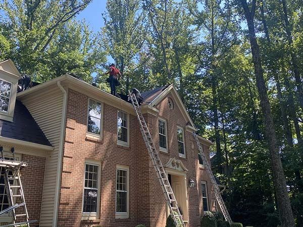 Workers on ladders repairing a brick house roof amid tall trees