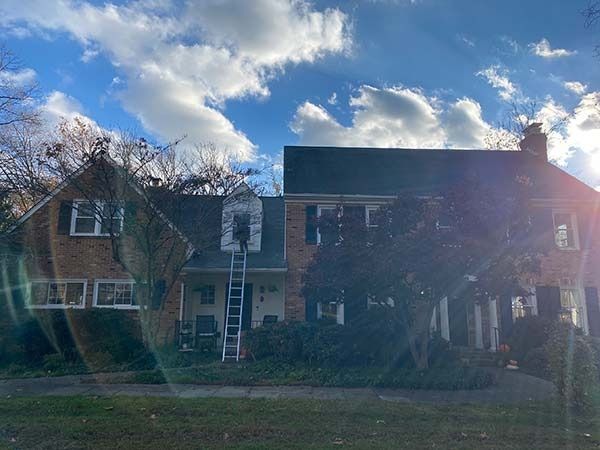 Brick townhouse with white ladder on front, partly obscured by trees, under a cloudy blue sky