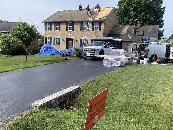 Workers on a suburban roof with trucks, supplies, and tarps beside a driveway and lawn