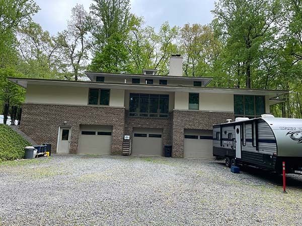 Brick house with three garage doors and a parked RV on a gravel driveway, surrounded by trees.