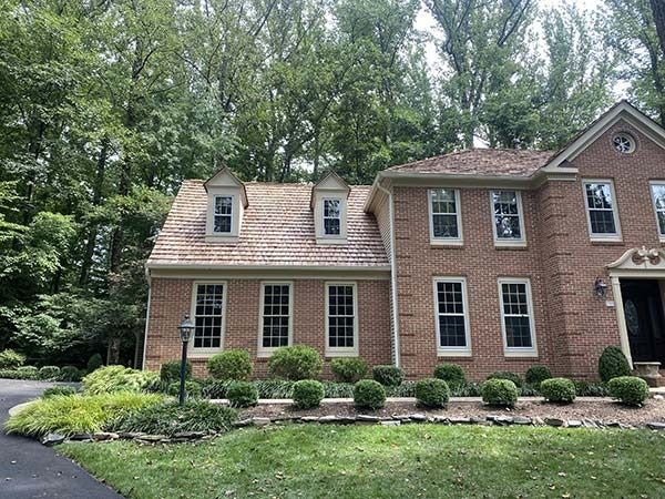Brick house with dormer windows and manicured shrubs beside a wooded driveway