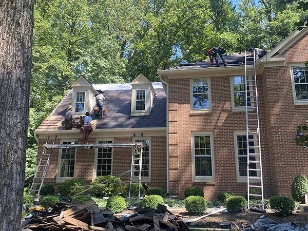 House with damaged roof and workers repairing it amid debris and trees.