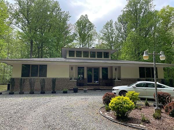 Single-story house with stone columns, gravel driveway, and parked white car surrounded by trees.