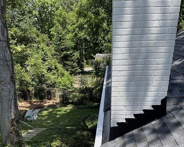 Wooded backyard beside a gray house roof and chimney, with lawn chairs on a grassy slope.