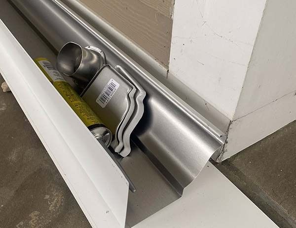 Metal duct and vent parts on a floor beside a wall, with a white panel leaning nearby.