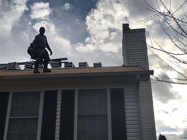 Person on a roof installing shingles beside a chimney under a cloudy sky