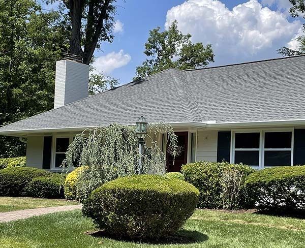 Single-story house with gray roof, white trim, and manicured shrubs in a sunny yard