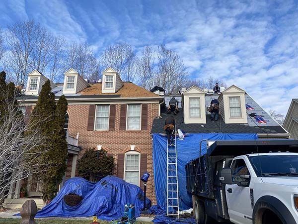 Brick house with blue tarps on roof and vehicles, with a ladder leaning up the front.