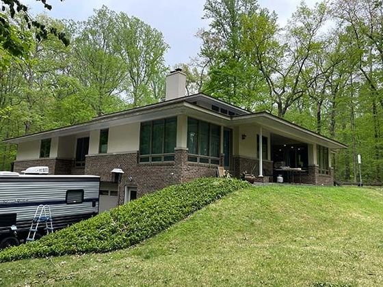 Single-story brick house with green shutters on a sloped lawn, surrounded by trees.