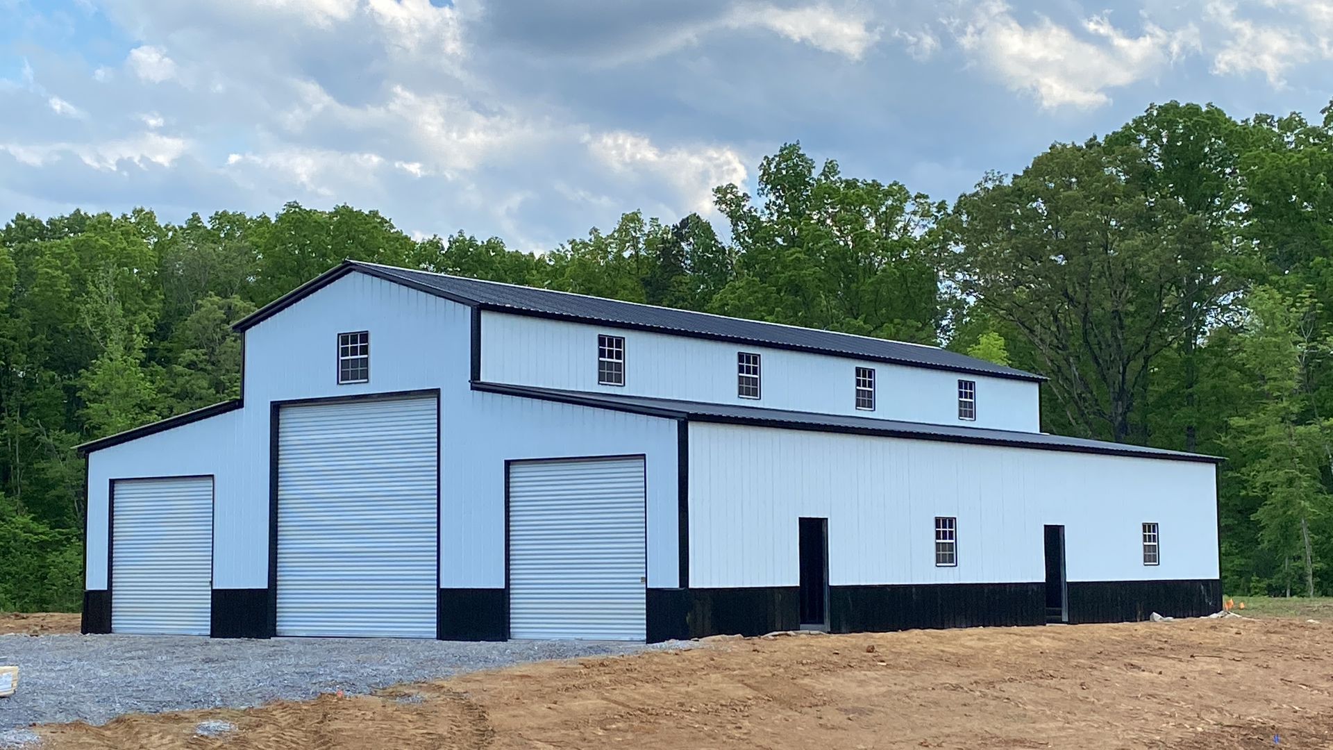 Large white metal barn or workshop with black trim and garage doors, set on a gravel lot by trees and cloudy sky