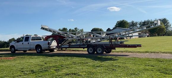 White pickup truck towing a red car hauler trailer on a grassy field under a blue sky