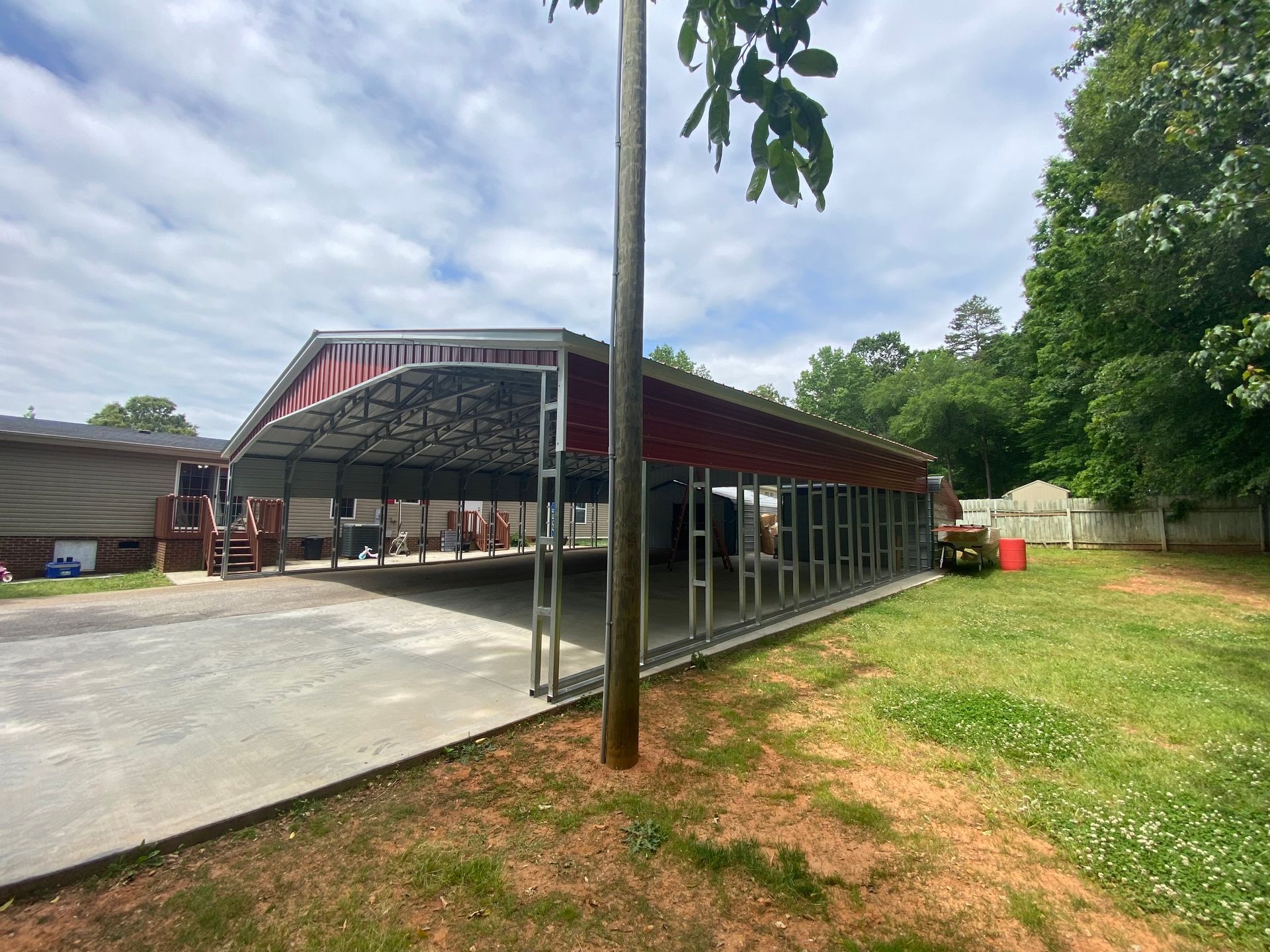 Covered outdoor pavilion with red roof, concrete floor, and grassy yard beside a tall pole and trees.