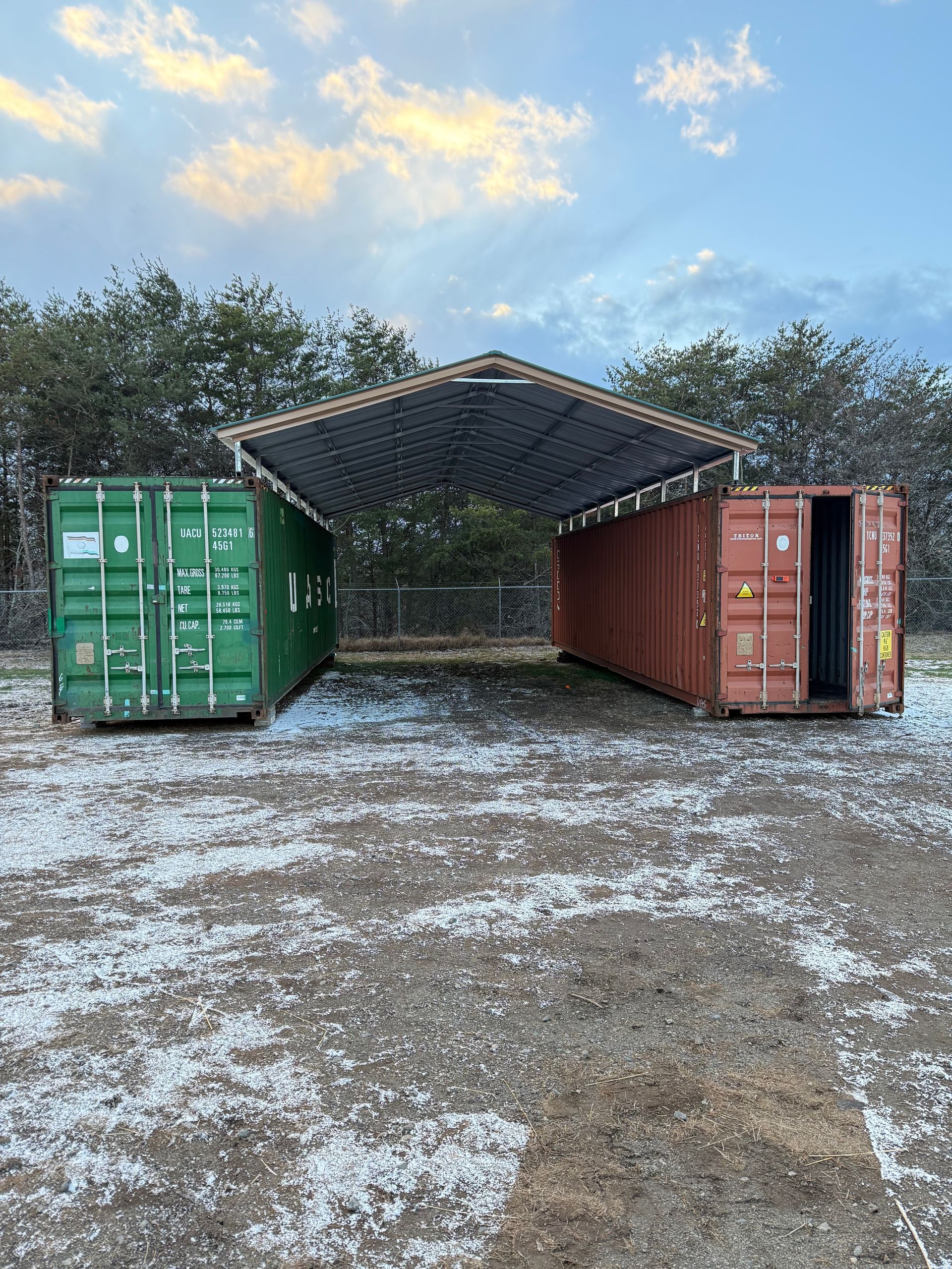 Two shipping containers, green and rust-red, with a metal roof canopy on a snowy gravel lot