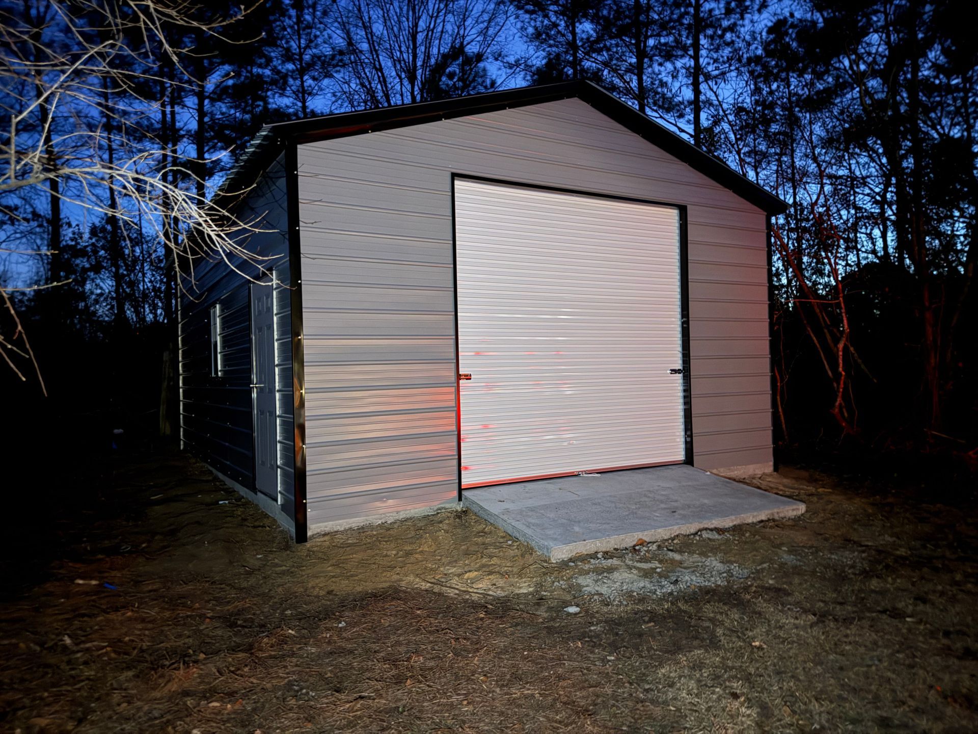 Small gray shed with a white roll-up door at dusk, surrounded by trees.