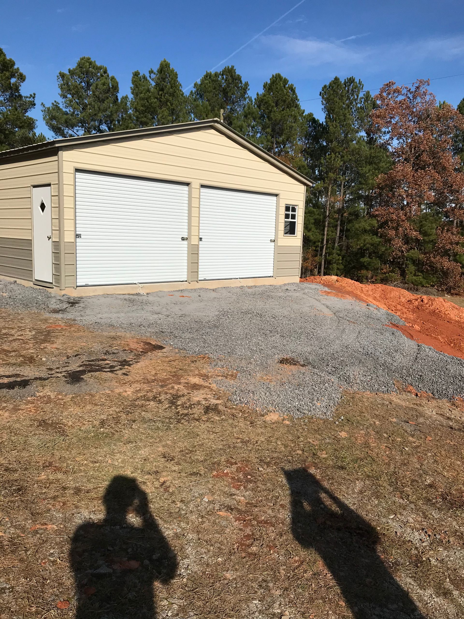 Detached tan garage with white double doors on a gravel driveway, trees and blue sky behind it.