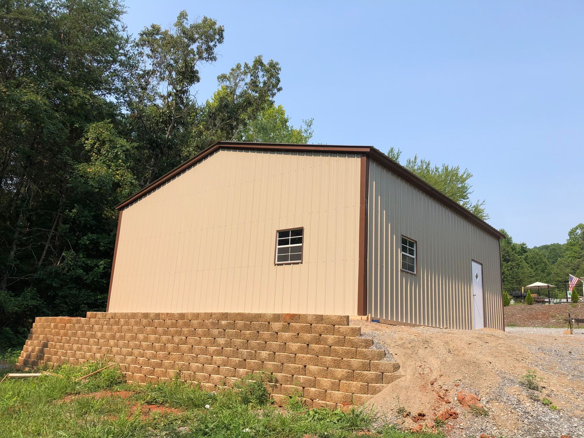 Tan metal-sided building on a gravel lot, with a stacked-stone retaining wall and trees in the background.