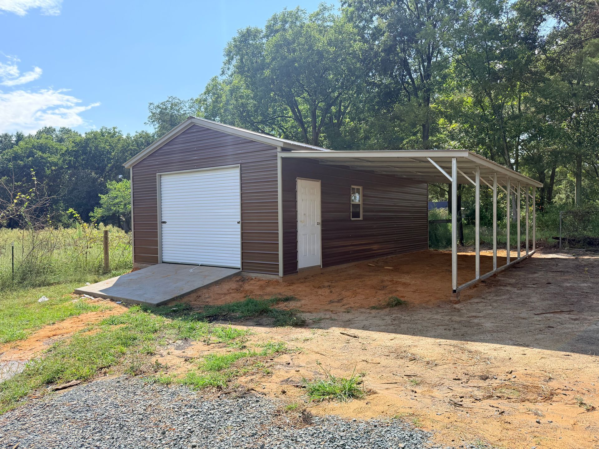 Small brown garage with white door and covered side porch on a dirt lot, surrounded by trees