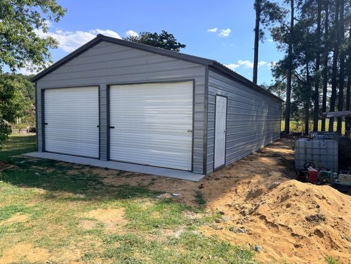 Gray metal garage with two white roll-up doors, shown on a dirt lot with trees in the background.