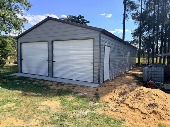 Gray metal garage with two white roll-up doors, set on a dirt lot beside a wooded area.