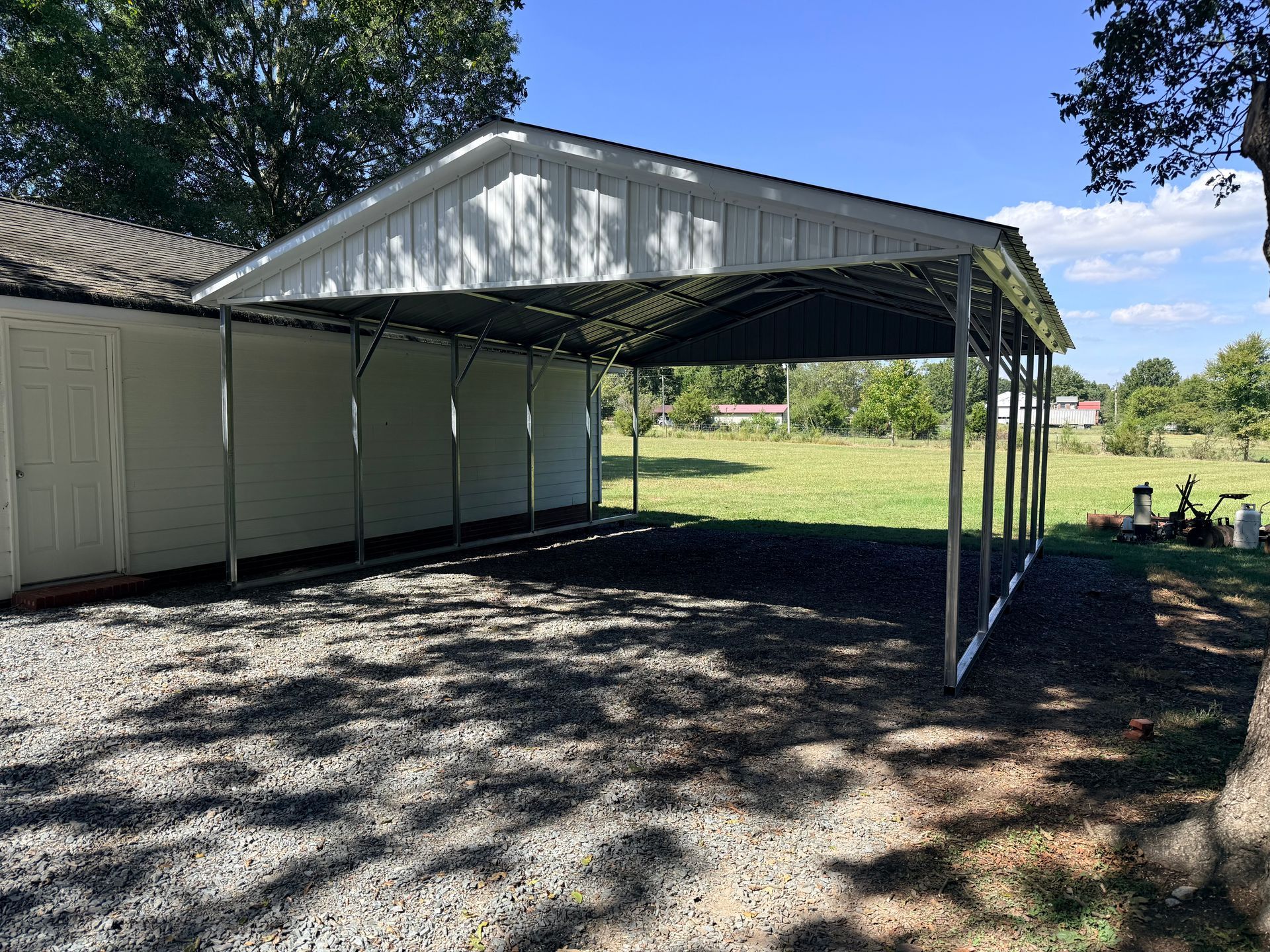 White barn with a metal-roofed carport on gravel beside a grassy yard under trees